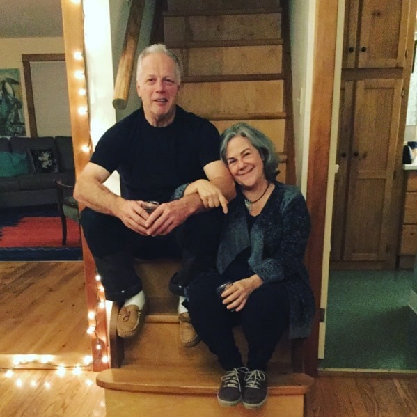 Corrine Koslo in a grey-black cardigan and black pants posing with her husband in a black t-shirt and pants.